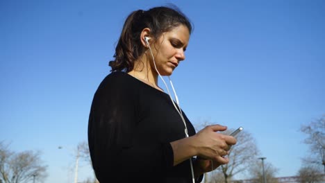 Focused-young-woman-in-earphones-using-smartphone-in-park
